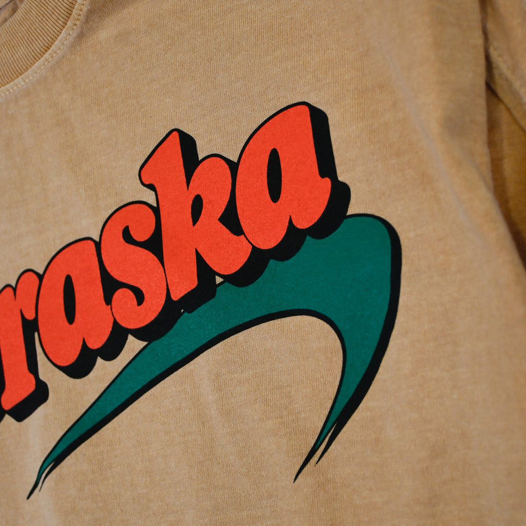 Print detail of orange Nebraska lettering and green graphic with black shadow on tan tee