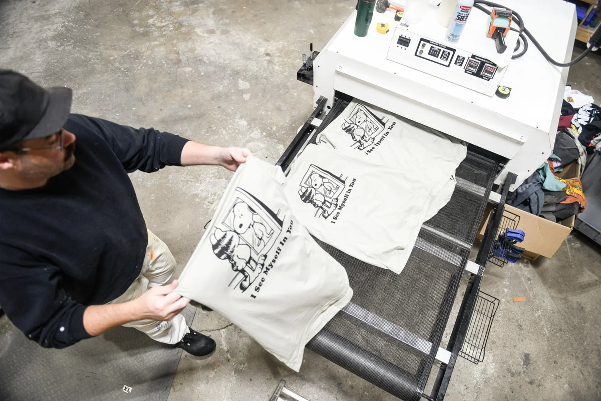 Omaha Screen Co printer handling freshly printed cream T-shirts beside the conveyor dryer